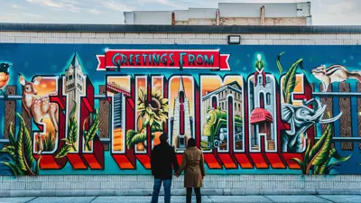 Two people stand in front of a colourful mural that says "Greetings from St. Thomas"